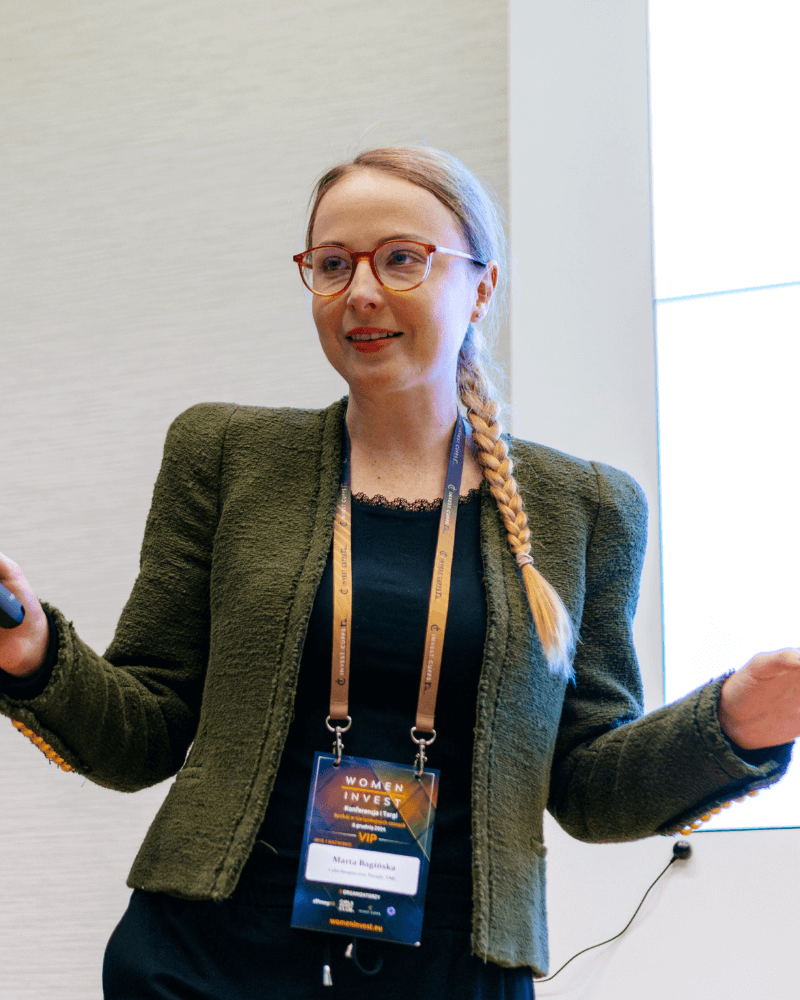 Marta Bagińska Cyberbezpieczne (2) (1) Marta Bagińska w trakcie wystąpienia na konferencji. Ma na sobie zieloną marynarkę i plakietkę konferencyjną na szyi.