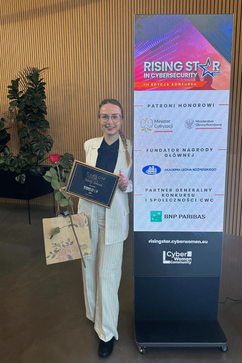 Marta Bagińska - cyberbezpieczne (1) Marta Bagińska - w rękach trzyma kwiaty, dyplom nagrody Rising Star in Cybersecurity i torebkę prezentową. Uśmiecha się.